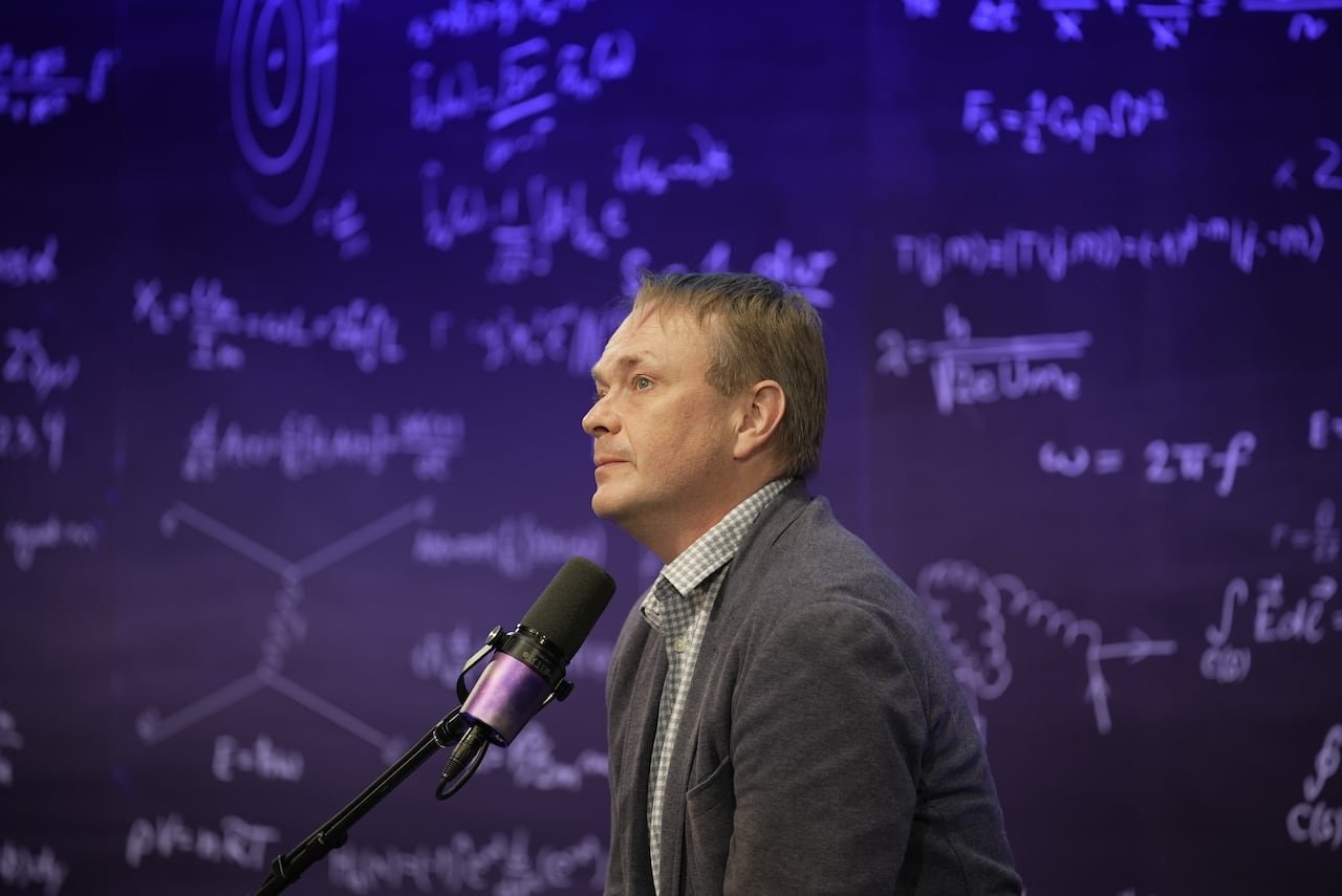 default-335 A man wearing a dark gray suit sits in front of a podcast microphone.