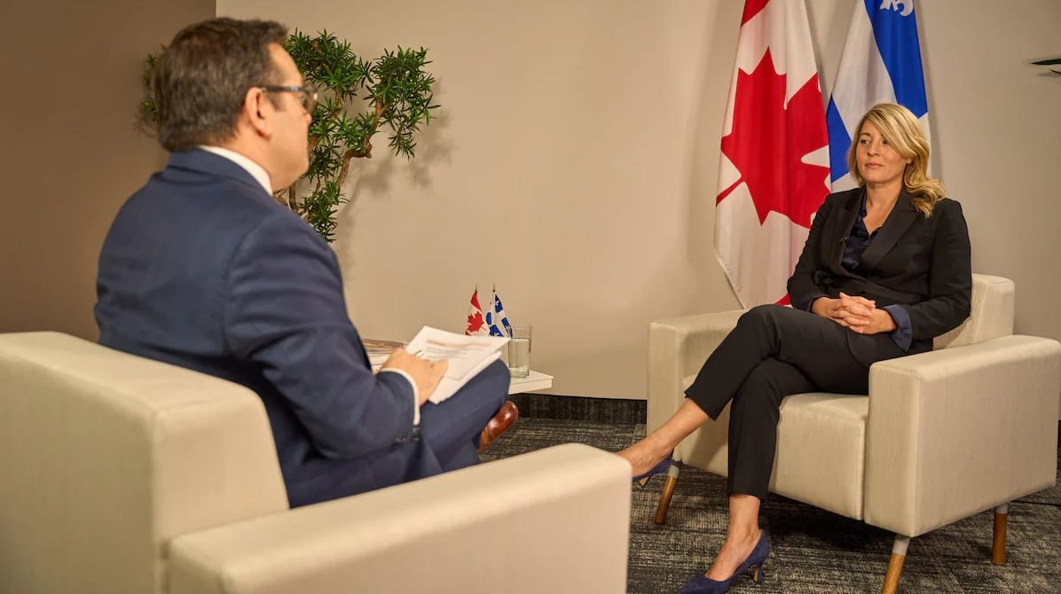 default-912 A woman in a suit sits in a chair in front of a Canadian and Quebec flag. A man in a suit sits across from her facing away from the camera.