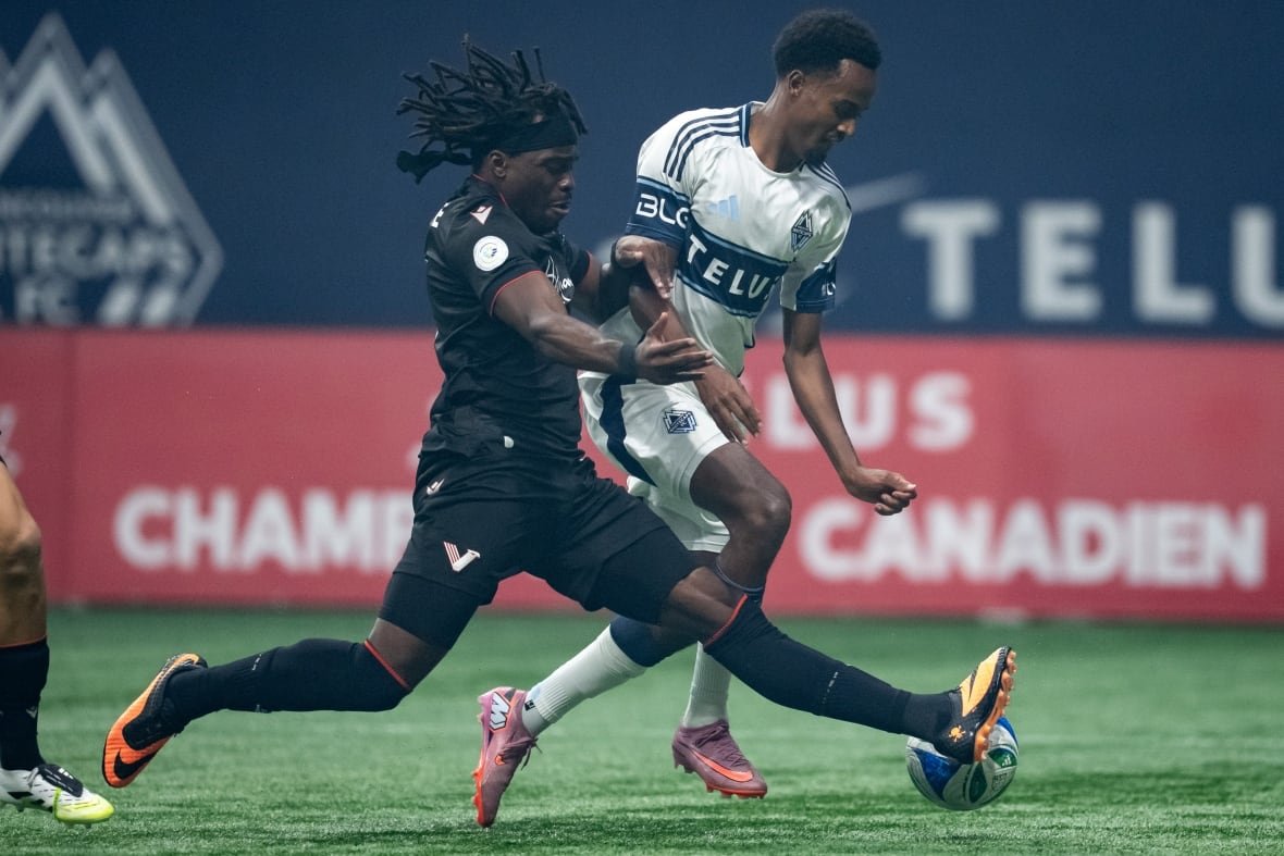 soc-vancouver-fc-whitecaps-20251001-2 Two soccer players, one in a black uniform and the other in a white and blue uniform, compete for a soccer ball.