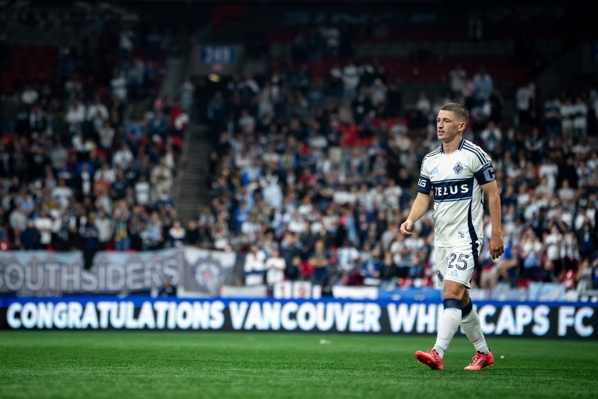 soc-vancouver-fc-whitecaps-20251001-4 Ryan Gauld wearing orange football boots and a white and blue uniform walking on the pitch at B.C. Place with fans in the background.