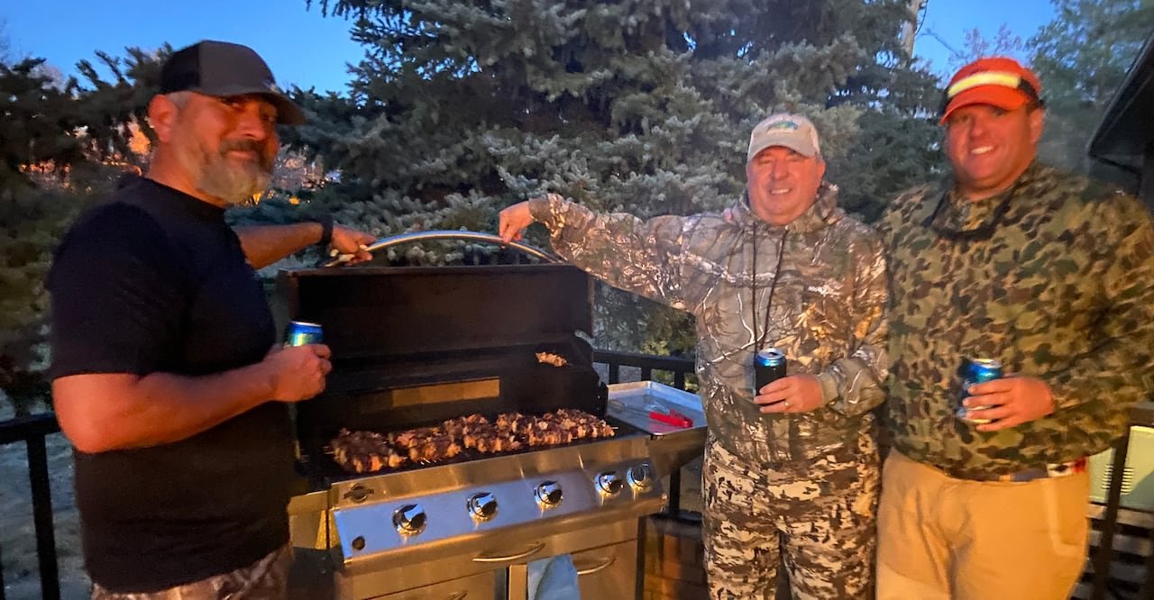 Three individuals in camouflage attire gather around a stainless steel barbecue grill preparing food.