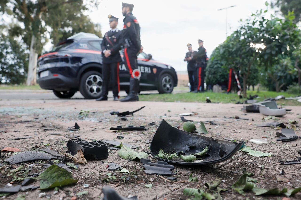 Debris from an exploded car scattered on the ground. Carabinieri military police officers stand near a police vehicle.