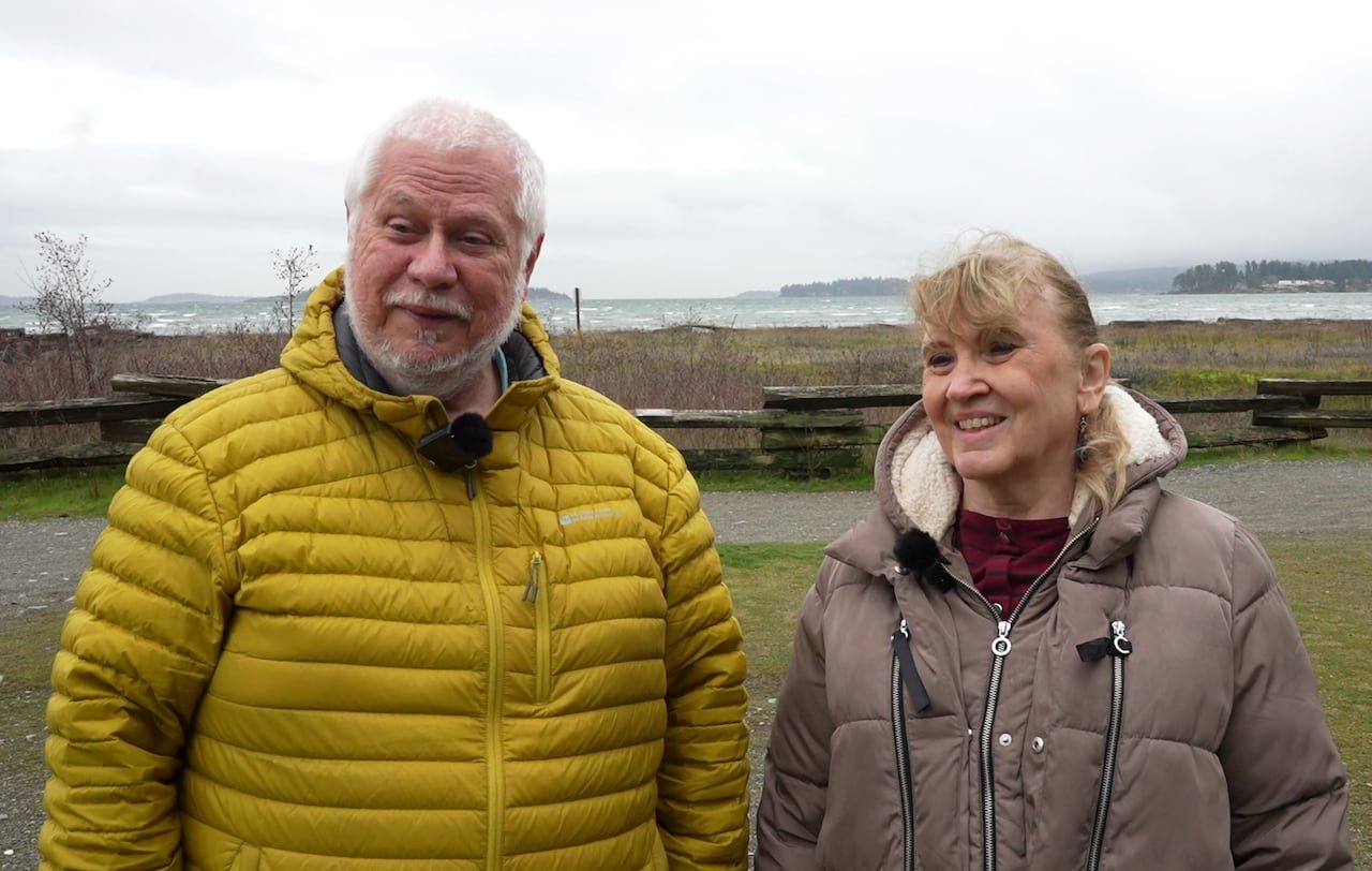 A man in a yellow jacket and a woman in a brown jacket smile outdoors.