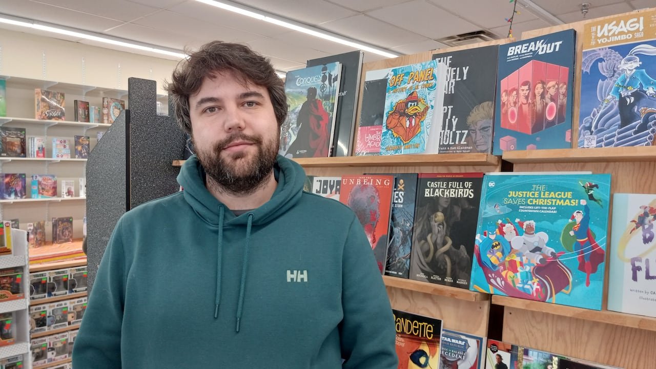 default-429 Man in teal sweater standing next to a bookshelf filled with comic books.