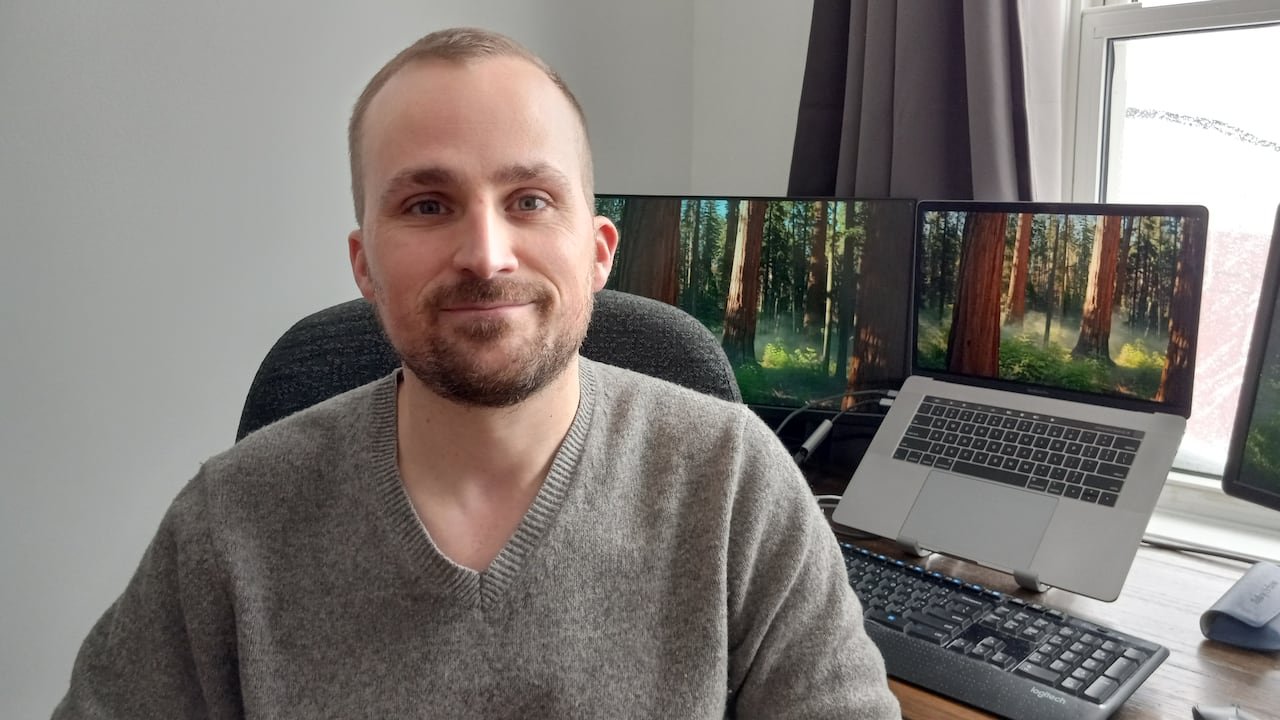 default-430 Man in grey sweater sitting at a desk with two laptops behind him.