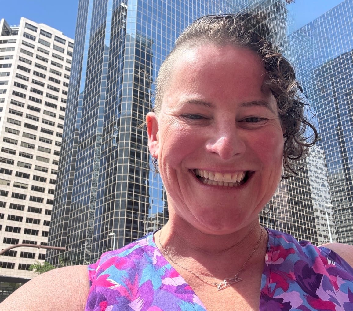 default-784 A woman with short curly hair shaved on one side smiles broadly with tall office buildings in the background.