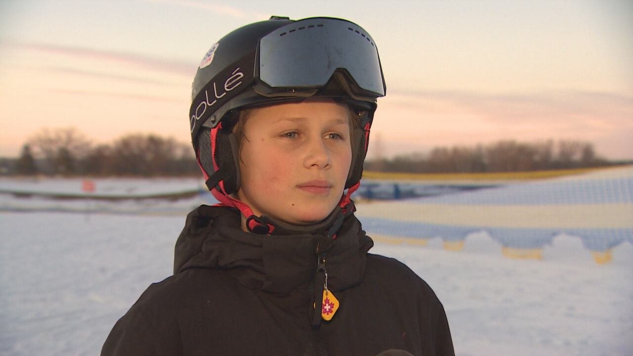 A boy in ski gear stands outdoors near a ski slope.