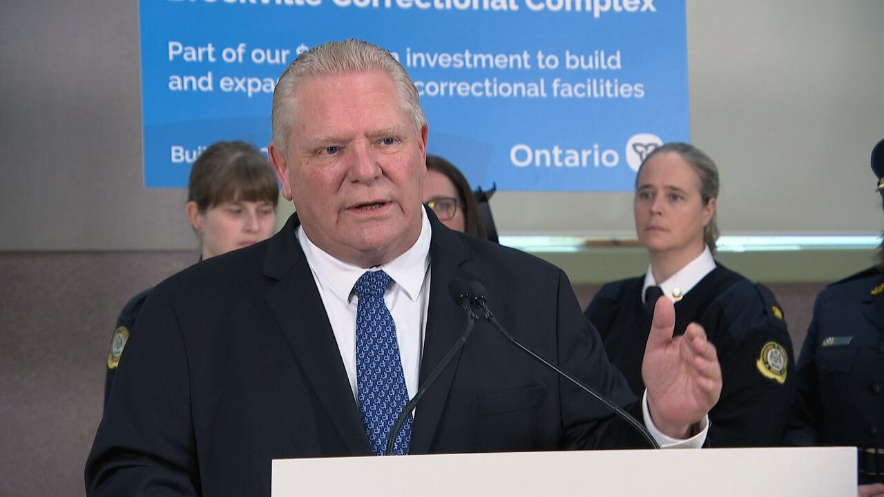 default-249 A man with short blonde hair, wearing a suit and blue tie stands in front of a podium. Behind him are jail guards in uniform.