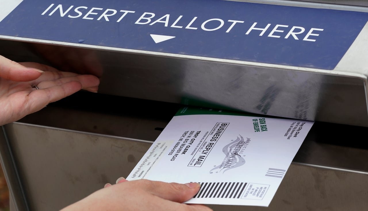 A hand holds a mail-in ballot envelope at the opening of a drop box labelled 'Insert Ballot Here'