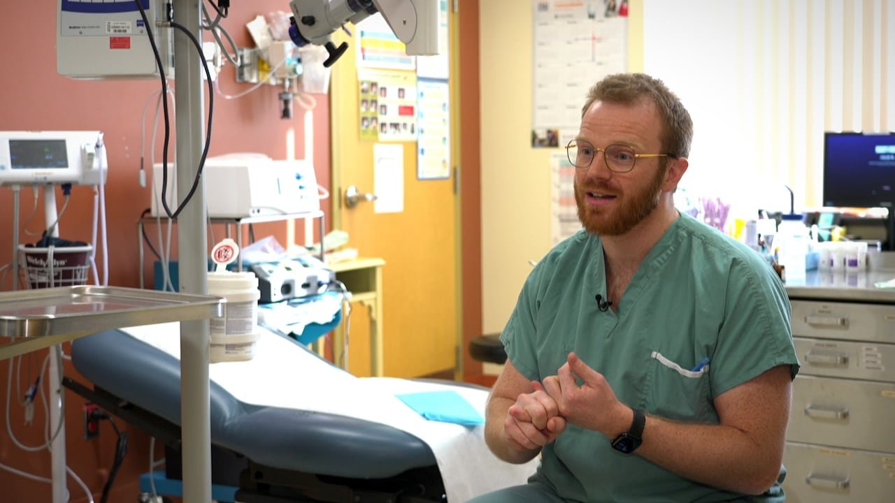 A man wearing scrubs, sitting in an exam room