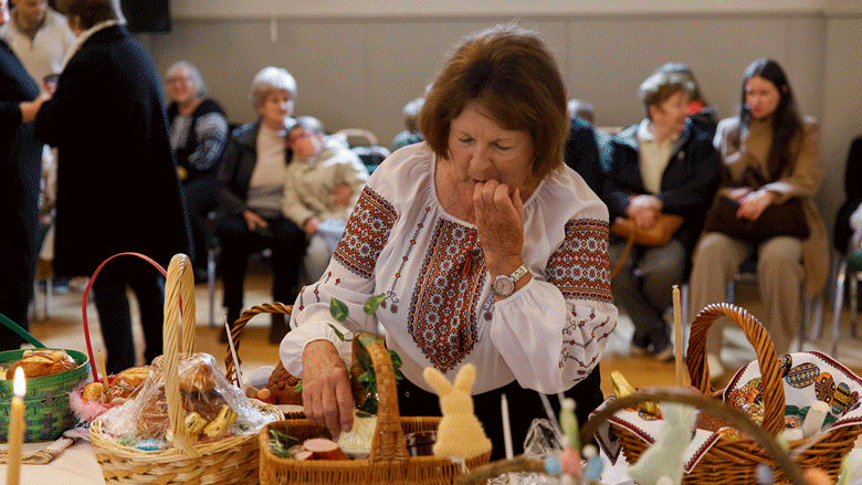 default A woman at a table filled with food baskets, gazing at a basket, and a close-up of bread, eggs, treats, sausages, and candles.